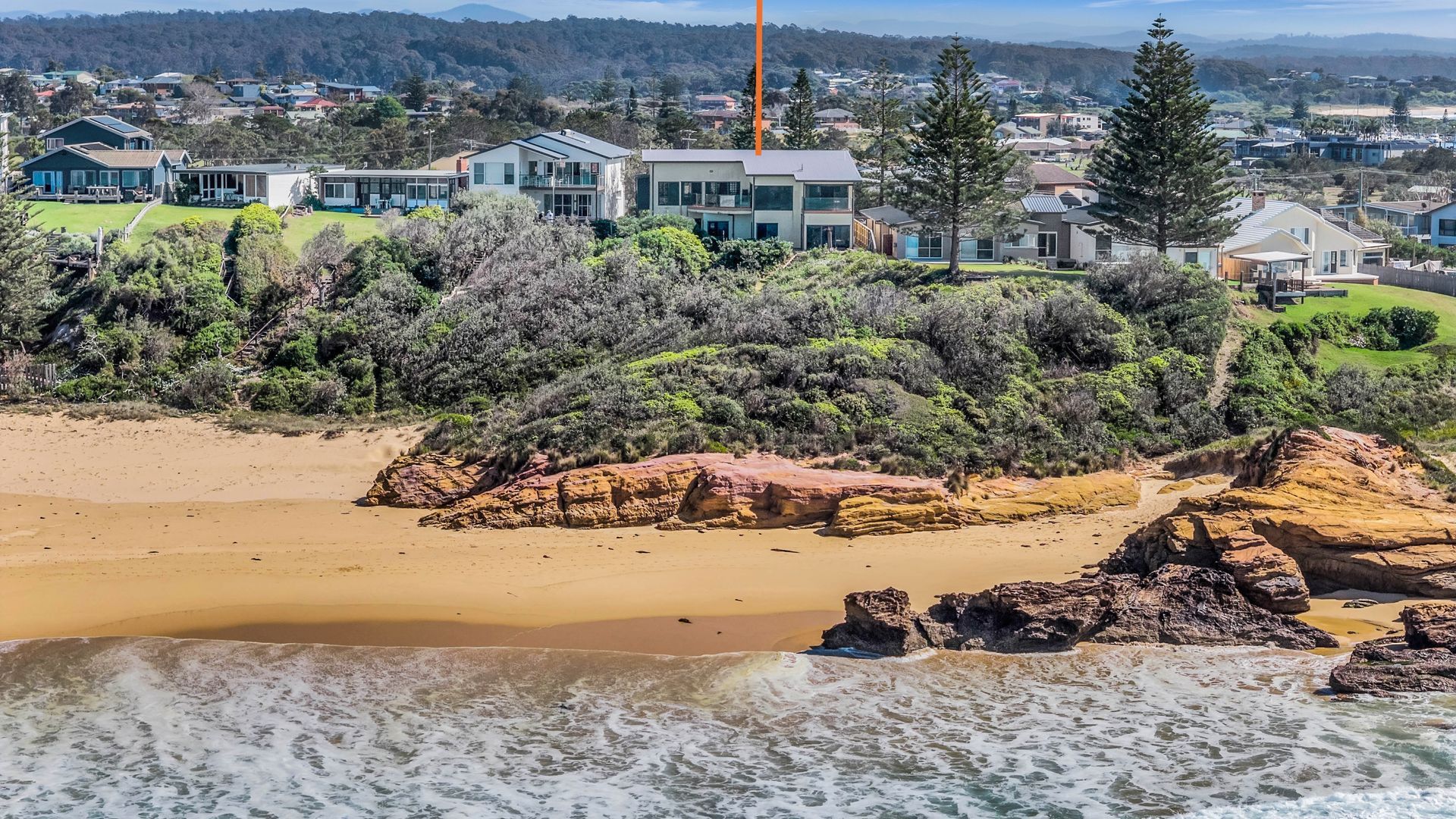 A coastal property with a sandy beach, rocky outcrops, and a backdrop of residential houses.