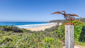 A coastal landscape with a metal sculpture on a wooden post, overlooking a beach and ocean.