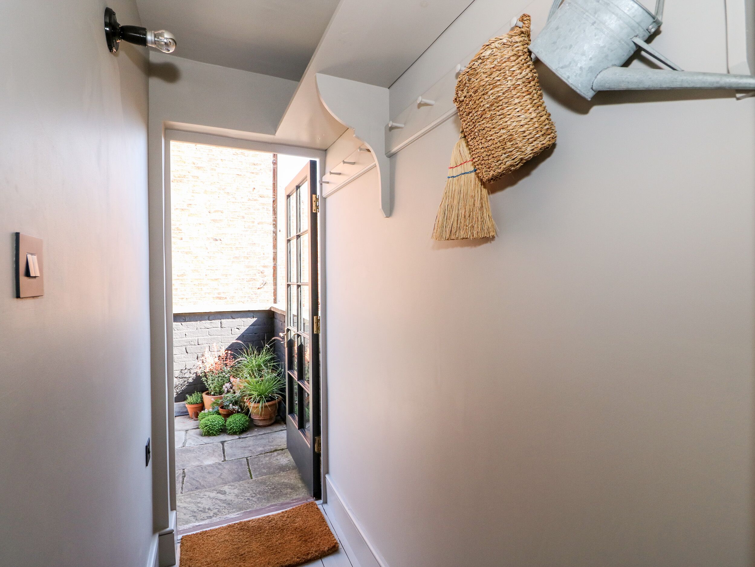 A narrow hallway with a door leading to a brick exterior. There are potted plants and a small rug at the entrance.