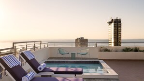 A rooftop pool area with lounge chairs and a table, overlooking the ocean and a tall building. - Home Suite Hotels Sea Point (Cape Town)