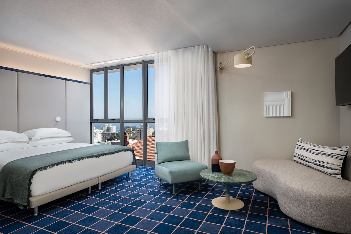 Et hotelværelse med seng, sofa, stol, et lille bord og et stort vindue med udsigt over byen.