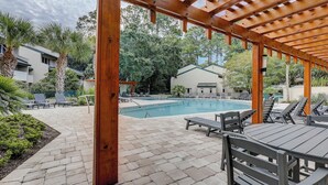A covered outdoor pool area with lounge chairs and a table, surrounded by greenery and buildings. - 256 Stoney Creek - Walk to Harbour Town to watch the Pros at the Heritage! (Hilton Head Island)