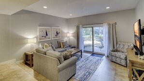 A spacious living room with a large sofa, a smaller sofa, a coffee table, and a TV. There are two wall-mounted lamps and a floor lamp. The room has a large sliding glass door leading to an outdoor area. - 256 Stoney Creek - Walk to Harbour Town to watch the Pros at the Heritage! (Hilton Head Island)