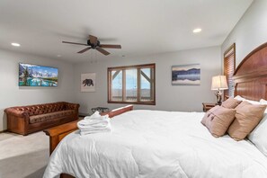 A bedroom with a large bed, a ceiling fan, a window with blinds, and artwork on the walls.