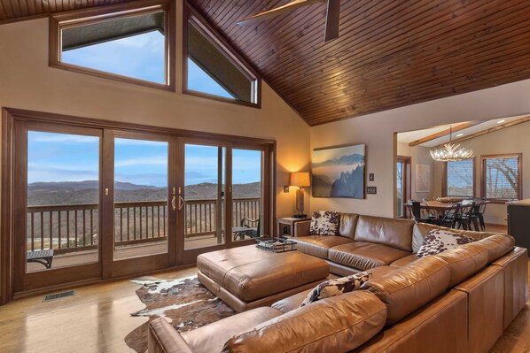 A spacious living room with a large leather sofa, a wooden ceiling, and a view of the mountains through large sliding glass doors.