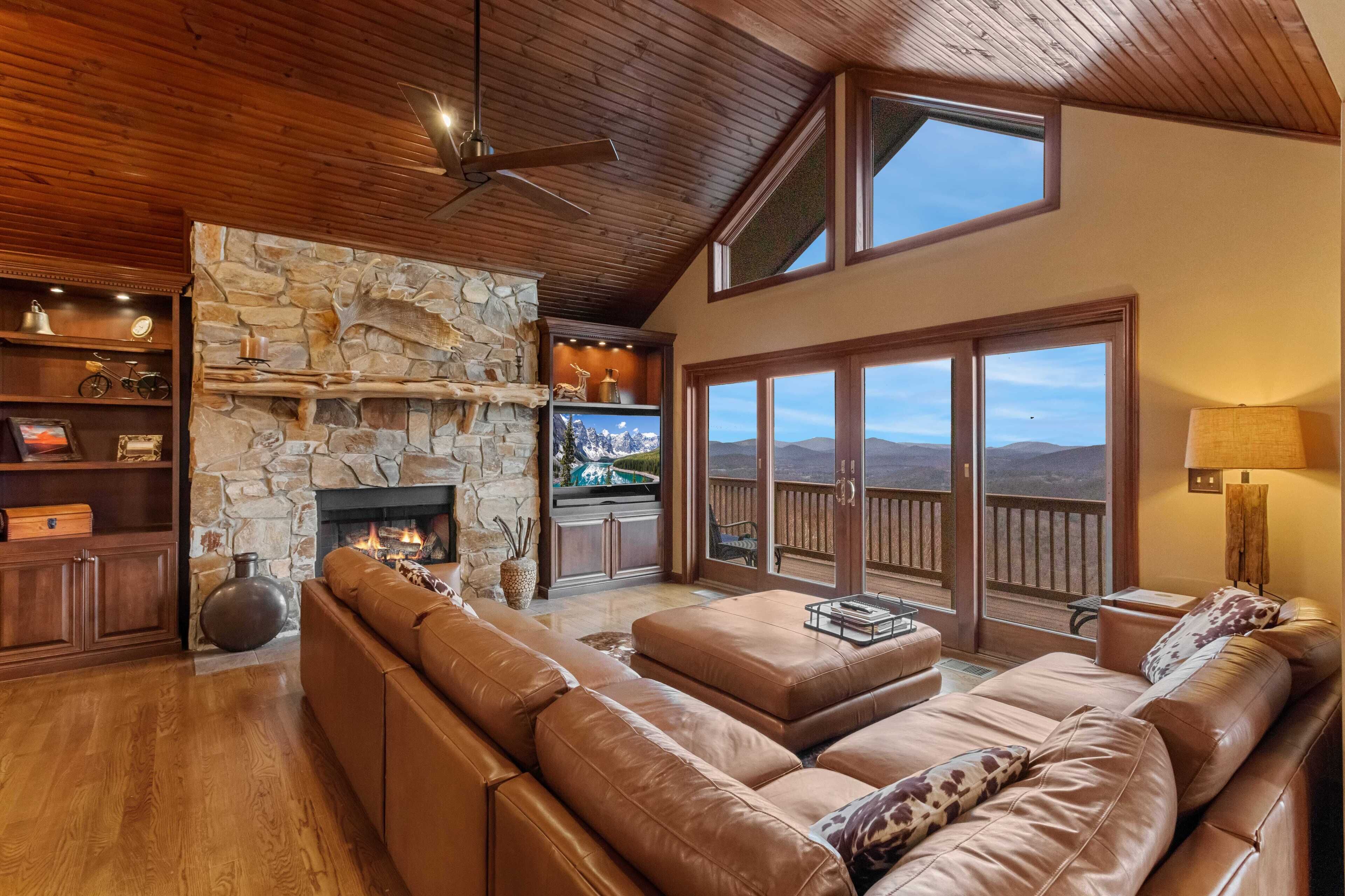 A spacious living room with a stone fireplace, leather sofas, wooden shelves, and a large window with a mountain view.