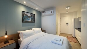 Hotellihuone, jossa on sänky, yöpöydät, seinään asennettu ilmastointilaite, ovi ja pieni keittiönurkkaus.