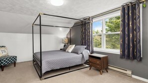 Un lit à baldaquin avec une couvre-lit grise, une table de chevet avec une lampe, et une vue sur la verdure à travers une fenêtre ornée de rideaux à motifs.