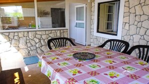A stone-walled room with a table covered in a floral tablecloth, two black chairs, and a window with a view of a kitchen. - 2 Bed Flat - Sleeps 8 - Parking - Wifi (Novalja)