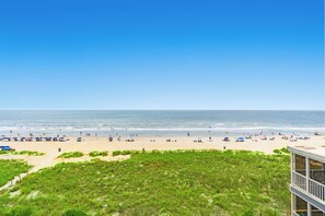 A beach with people and umbrellas, a clear sky, and a building with a balcony. - Charming 3 bedroom oceanfront condo in Crescent Beach | Shoreham Towers I - 5-E (North Myrtle Beach)
