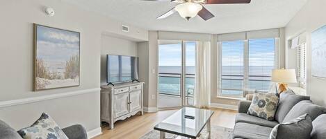 A spacious living room with a large window overlooking the ocean, a ceiling fan, and a television on a wooden cabinet.