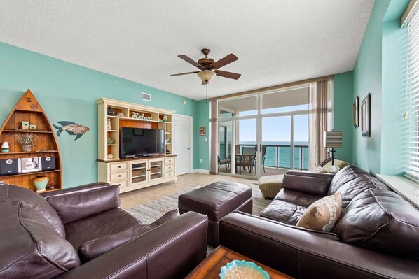 A living room with a television, a ceiling fan, a sofa, and a view of the ocean through large glass doors. - Luxurious Oceanfront Penthouse with Private Glass-Walled Balcony | Crescent Keyes - PH 9 (North Myrtle Beach)