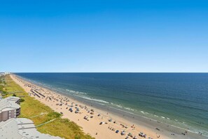 A beach with numerous umbrellas and people, a clear blue sky, and buildings along the shore. - Stunning condo with updates galore, oceanfront view | AshWorth - 1805 (North Myrtle Beach)