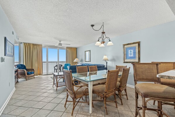 A spacious living area with a glass dining table surrounded by wicker chairs, a blue sofa, and a ceiling fan.