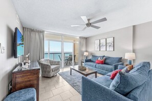 A modern living room with a flat-screen TV, a ceiling fan, and a view of the ocean through large sliding doors.