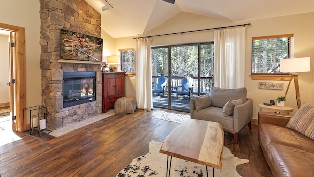 A spacious living room with a stone fireplace, a ceiling fan, a large window with curtains, and a leather sofa.