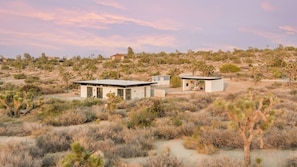 A modern house with a flat roof and large windows, surrounded by desert vegetation and sparse trees.