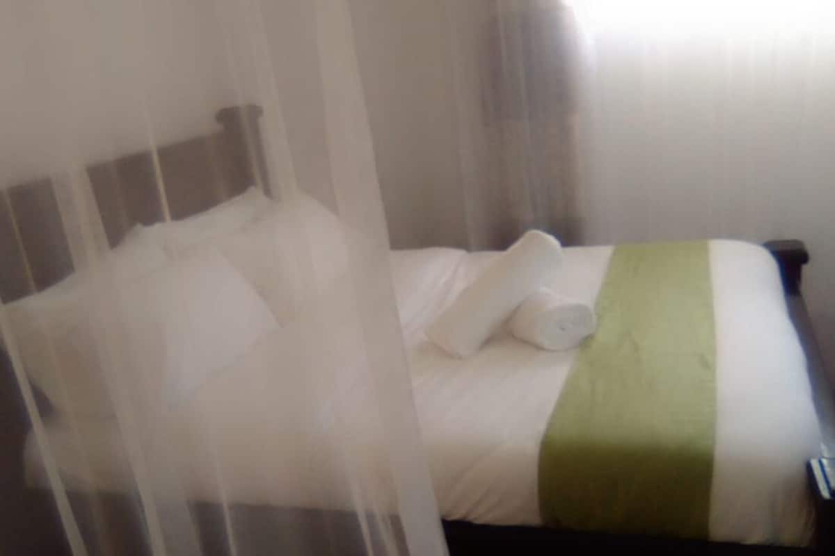 Un lit avec une literie blanche et une touche de vert, une tête de lit blanche et un rideau léger.