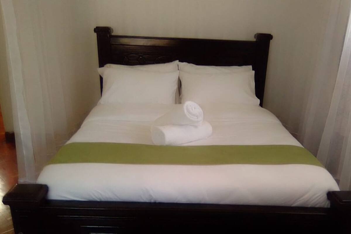 Un lit bien fait, avec du linge de lit blanc et une touche de vert, une tête de lit en bois foncé et des rideaux blancs.