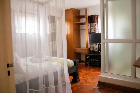 A bedroom with a bed, a wooden wardrobe, a small table, and a window with curtains. - Celio Jardine Lavington (Nairobi)
