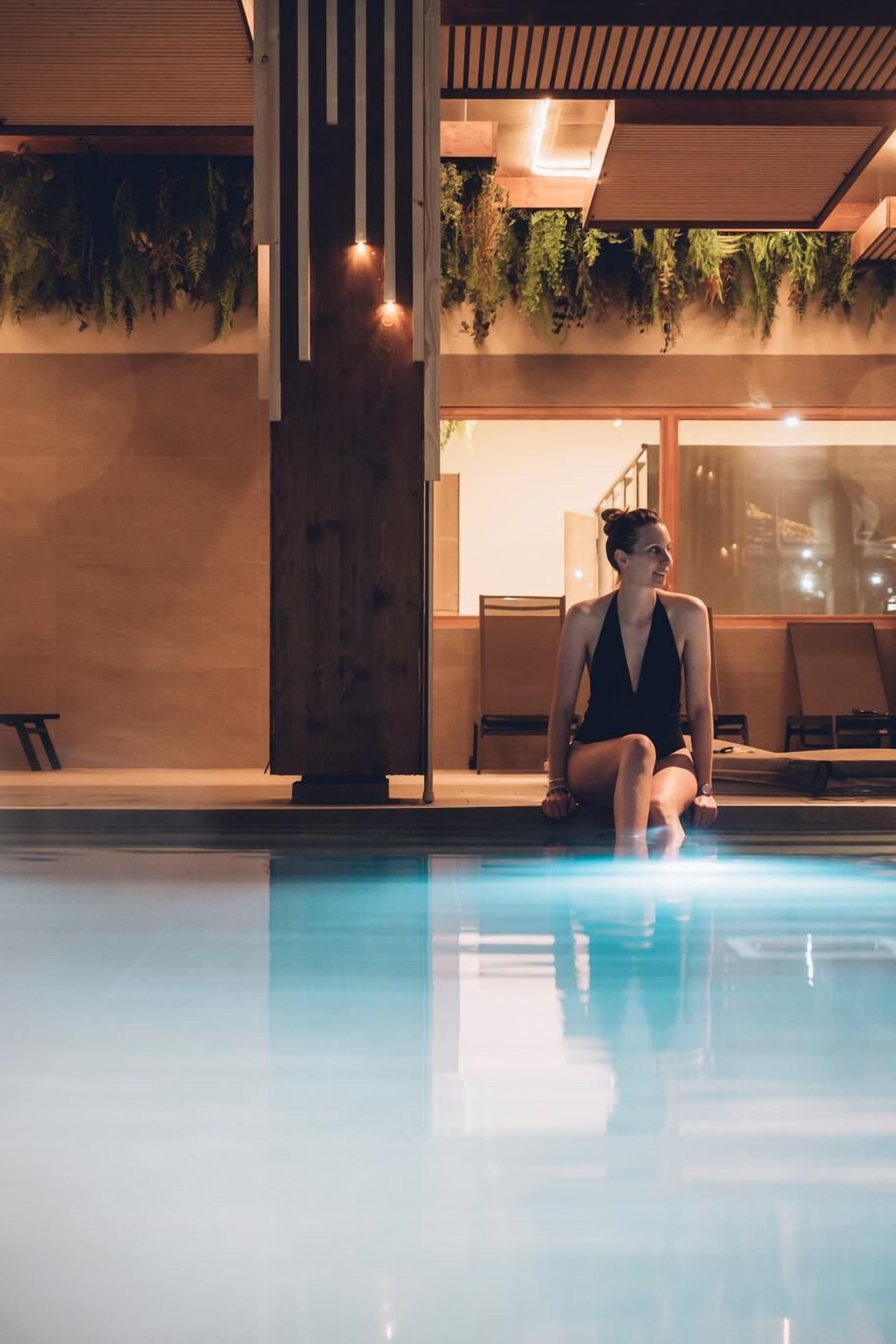 A woman sitting by an indoor pool with wooden pillars and hanging plants.