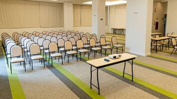 Uma sala de conferências com fileiras de cadeiras e mesas, piso coberto por carpete e uma grande janela.