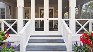 A white porch with a set of stairs leading to a door, flanked by flower beds with red and purple flowers. - Beach Access and Private Pool (Watersound)