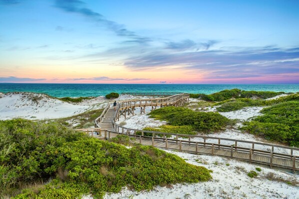 A wooden boardwalk leading to a beach with white sand and green vegetation.