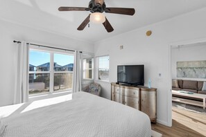 A bedroom with a large bed, a ceiling fan, a TV, a dresser, and a view of the outside.