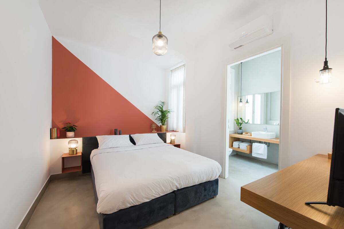 A modern bedroom with a bed, bedside tables, a nightstand, a desk, and a bathroom with a sink and mirror.