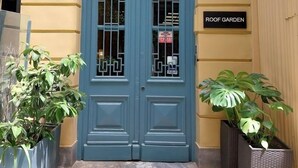 L’entrée d’un hôtel avec une porte bleue, une enseigne indiquant « MAISON DE MONK ATHÈNES – APPARTEMENTS » et une autre indiquant « JARDIN SUR LE TOIT ».