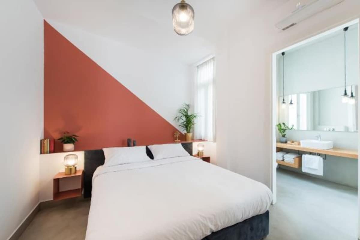 Una camera da letto moderna con un letto, comodini, una pianta in vaso e un bagno con lavandino e specchio.