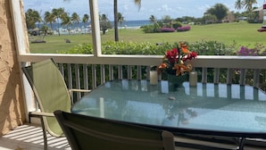 Ein Tisch mit Glasplatte und ein Stuhl auf einem Balkon mit Blick auf einen Strand, an dem Palmen wachsen und ein klarer Himmel zu sehen ist.