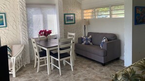 Ein Wohnzimmer mit Esstisch, weißen Stühlen, einem grauen Sofa mit gemusterten Kissen und einer Wand mit strukturierter Gestaltung.