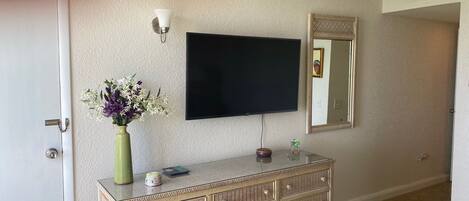 A room with a flat-screen TV mounted on the wall, a wicker console table with drawers, a vase with flowers, and a mirror.