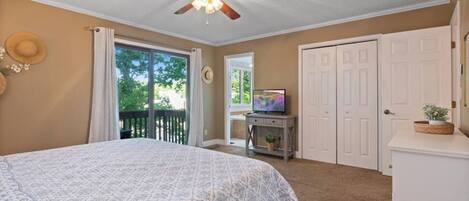 A bedroom with a large bed, a ceiling fan, a TV on a stand, and a sliding glass door leading to a balcony.