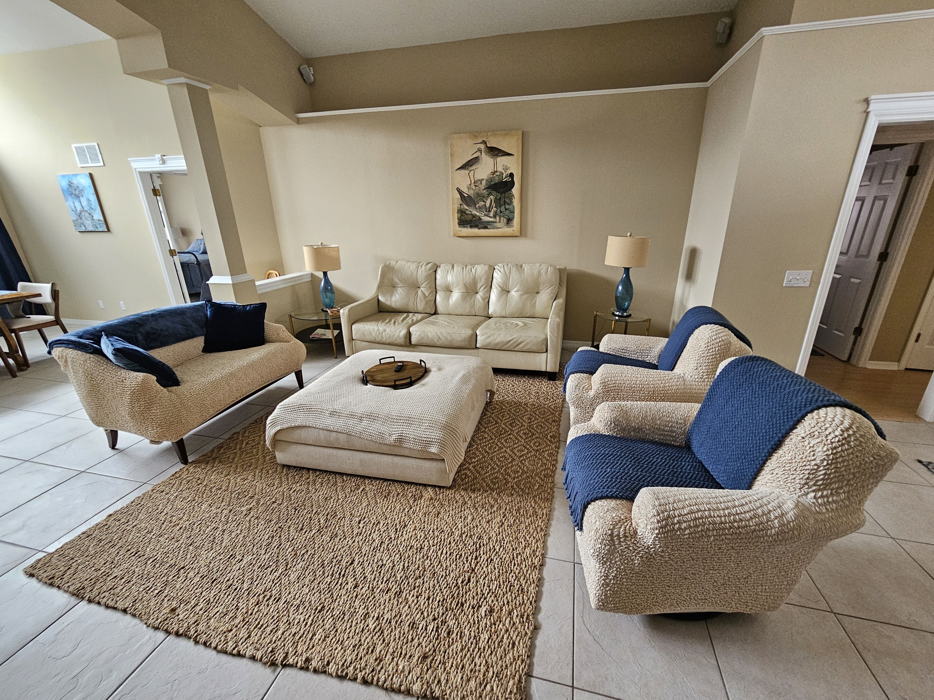 A living room with a beige sofa, a blue armchair, and a coffee table. There is a painting on the wall and a lamp on a side table.