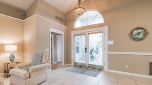 A spacious living room with a beige sofa, a chandelier, and a large window with a view of palm trees. - Peaceful Waterfront Pool Home 3 Bedroom Home by RedAwning (Palm Coast)