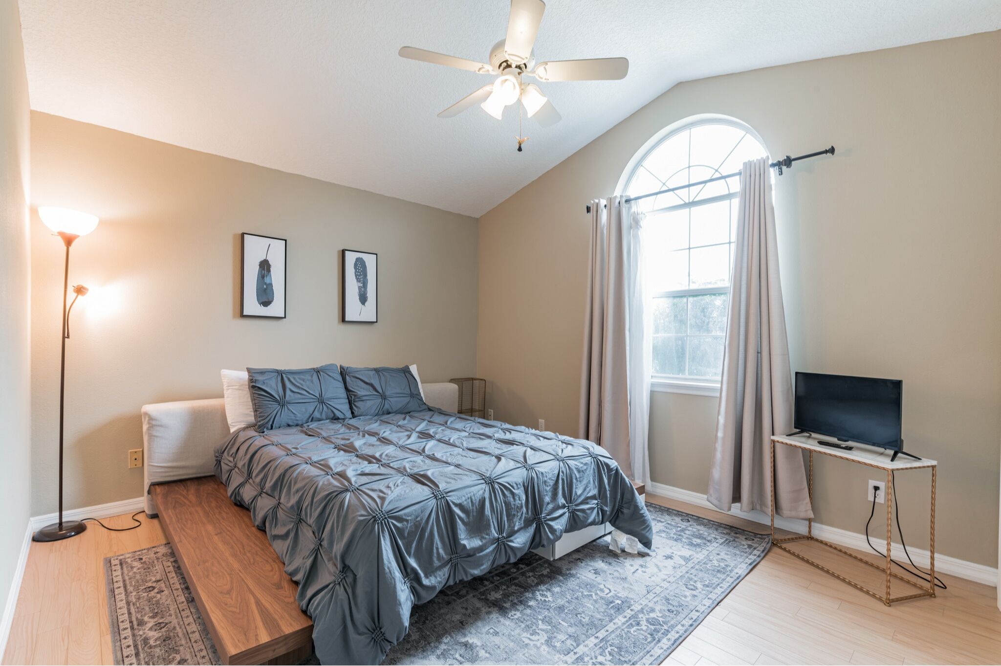 A bedroom with a bed, a standing lamp, a television on a stand, and a window with curtains.