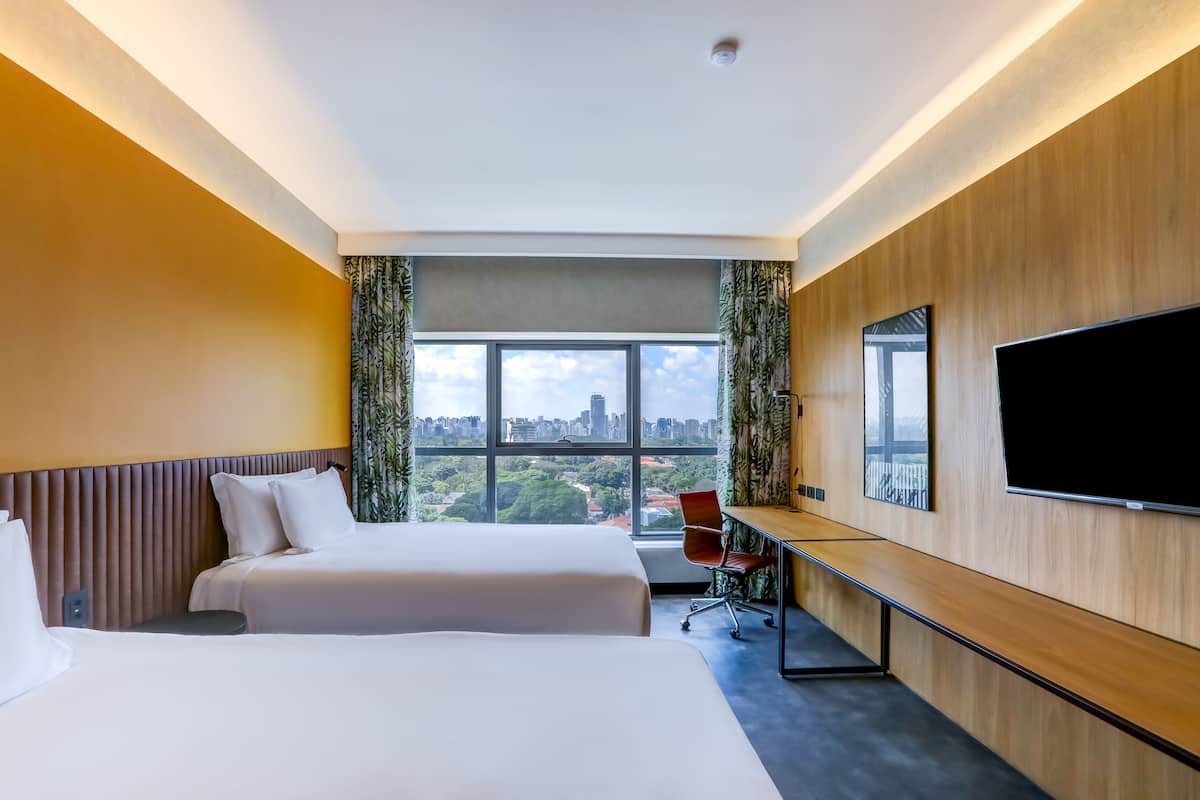 A hotel room with two beds, a desk, a chair, a TV, and a large window with a city view.