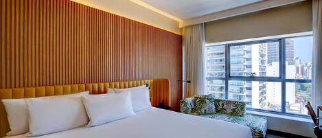 Habitación de hotel con una cama grande, cabecero de madera, vistas de la ciudad a través de un ventanal amplio y una pequeña zona de estar con un cojín estampado.