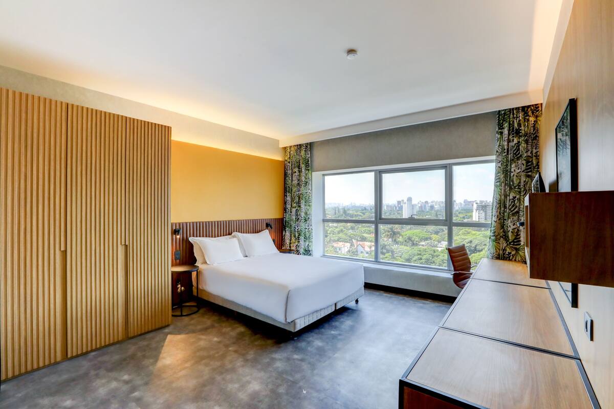A modern hotel room with a bed, a desk, and a large window offering a city view.