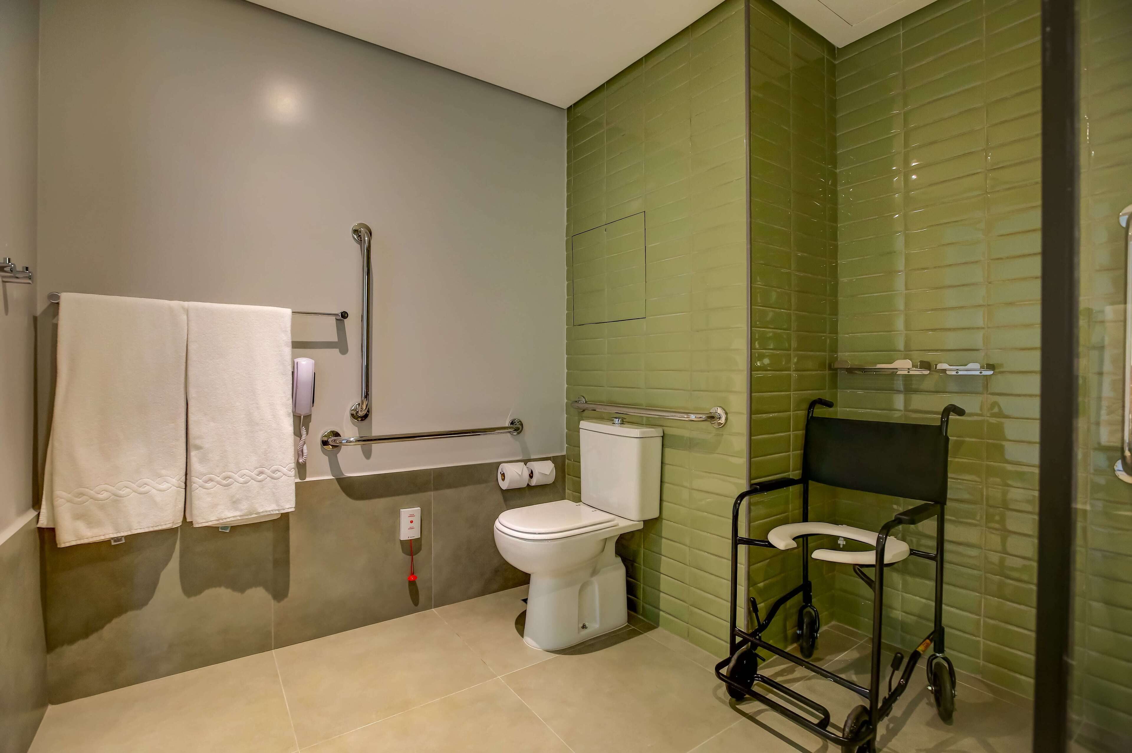 Une salle de bain équipée de toilettes, de barres d’appui, d’un fauteuil roulant et d’un porte-serviettes.