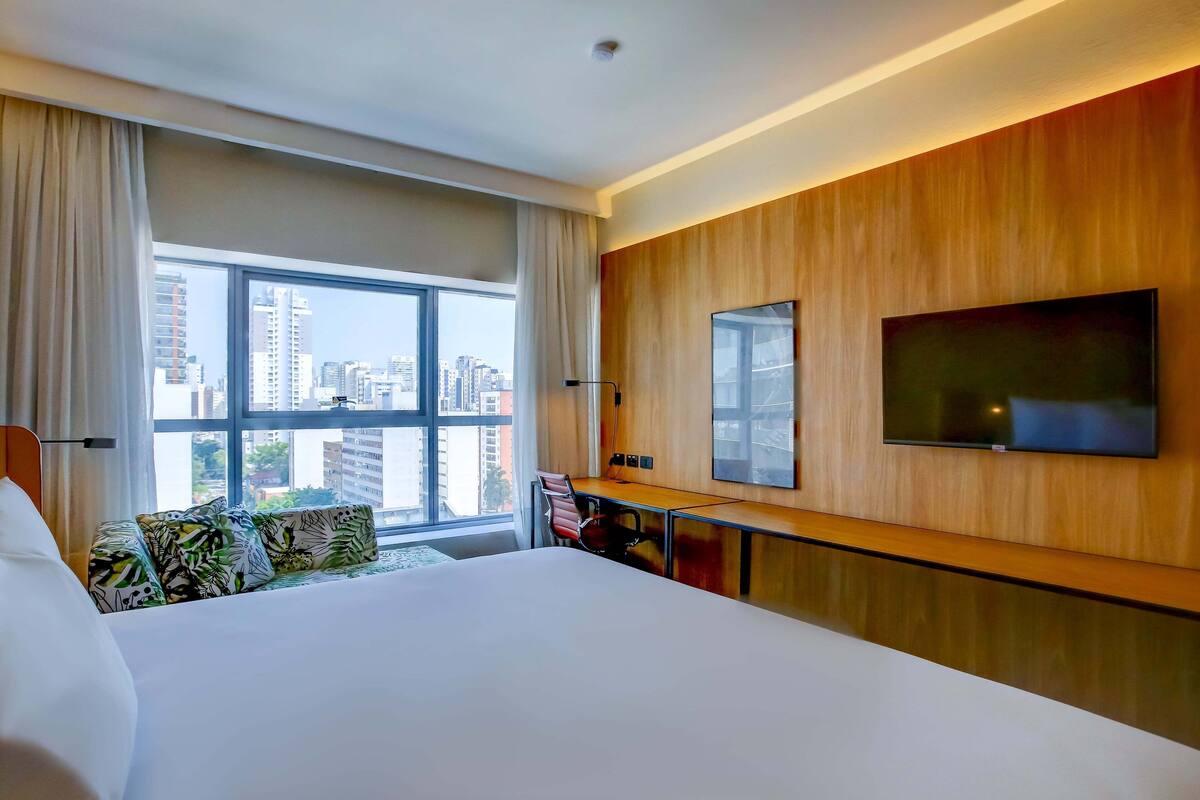 A hotel room with a large bed, a desk with a chair, a TV mounted on the wall, and a view of the city through a large window.