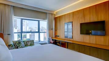 Habitación de hotel con una cama grande, un escritorio con una silla, un televisor montado en la pared y vistas de la ciudad a través de un amplio ventanal.