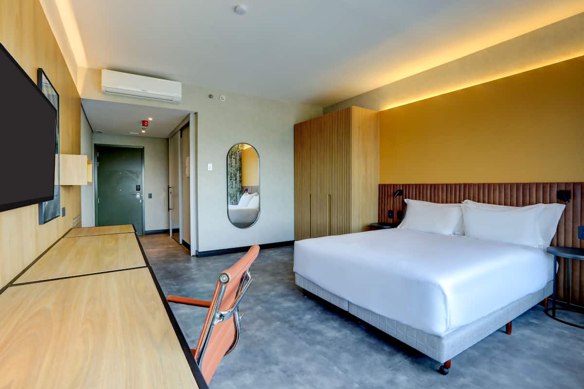 A hotel room with a large bed, a desk, a chair, a mirror, and a wall-mounted TV.