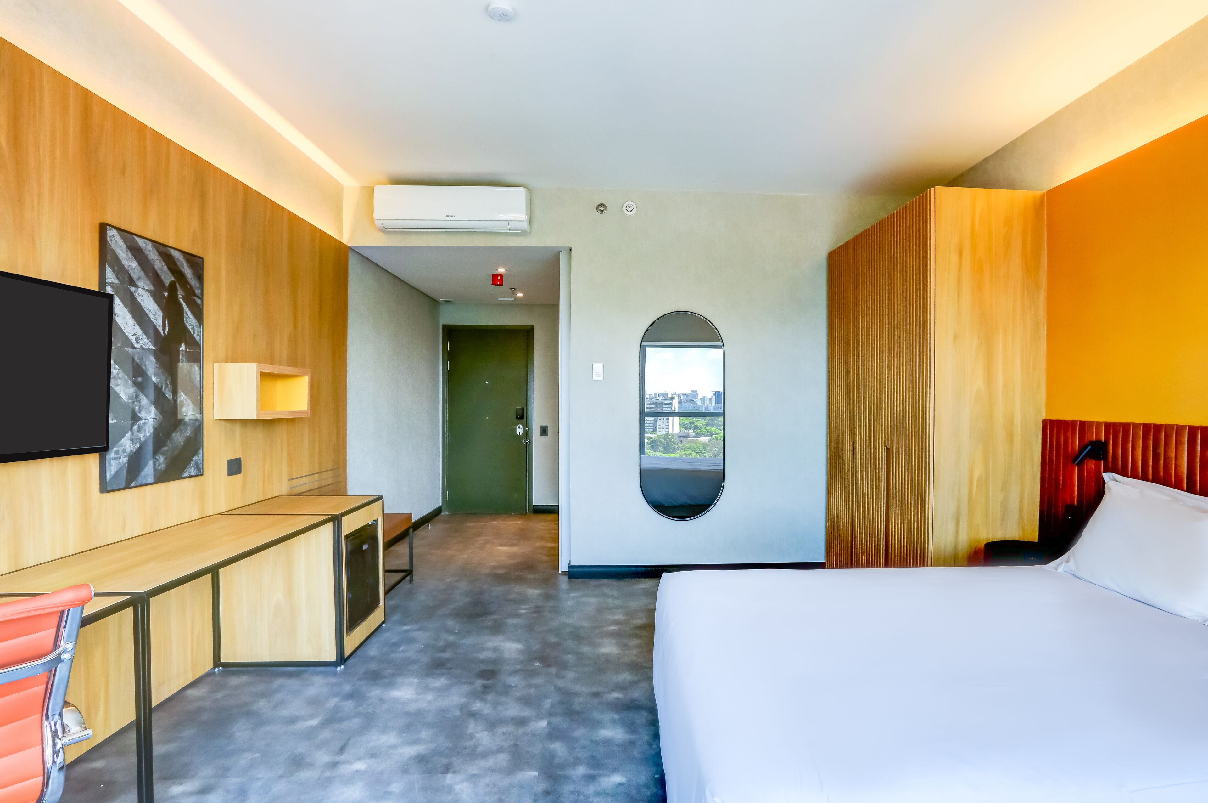 Quarto de hotel moderno com cama, escrivaninha e cadeira, vista para a cidade pela janela e ar condicionado.