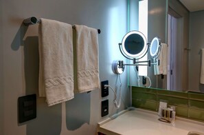A bathroom with two towels hanging on a towel rack, a round mirror with a light, and a small shelf with toiletries.