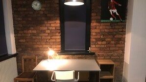 A room with a brick wall, a clock, a table with chairs, a lamp, and a poster. - Stylish Open Plan Loft in Manchester City Centre (Manchester)