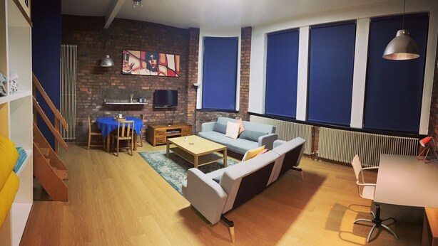 A living room with a sofa, coffee table, and TV. There is a staircase, a dining area with a table and chairs, and a radiator. - Stylish Open Plan Loft in Manchester City Centre (Manchester)